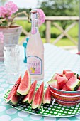 Green and white spotted plate, red striped bowls and sliced watermelon in front of retro lemonade bottle on table in summery garden