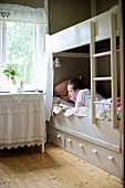 Teenager lying on white wooden bunk bed next to side table with valance below window