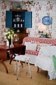 Various chairs, wooden table, sofa, crockery in open corner cabinet and floral wallpaper
