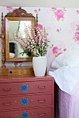 Vase of flowers and antique vanity mirror on mauve chest of drawers with pattern of blue flowers against floral wallpaper