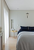 Doppelbett mit hellgrauer Decke, Krähenfuss Tisch (Traccia) zwischen Bett und bodenlangem Vorhang am Fenster