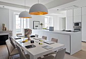 Open-plan, bright kitchen with modern table below pendant lamps with white and gold lampshades