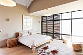 Animal-skin rug on fine wooden floor next to simple double bed in spacious bedroom with wooden, slatted sliding elements on glass wall