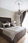 Box-spring bed with brown valance and stacks of scatter cushions against headboard in traditional bedroom with chandelier hanging from stucco ceiling rose