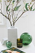 Branches of leaves in white ceramic vase and green drinking glass between two green glass spheres on white table