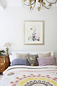 Double bed with patterned bedspread and many scatter cushions against headboard below modern artwork on wall