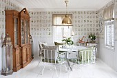 White metal chairs with cushions around round table and glass-fronted dresser in dining room with botanical wallpaper and white wooden floor