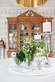 White flowers and lit candles in candlesticks on table in front of antique, glass-fronted cabinet
