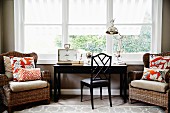 Wicker armchairs with seat cushions and scatter cushions flanking dark wood writing desk and chair below window with view of garden