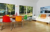 Colourful shell chairs and wooden table in front of panoramic window with woodland view; sofa with brightly striped scatter cushions to one side