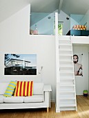 White couch with striped scatter cushions and large-format photographs flanking narrow, steep stairs leading to mezzanine under sloping ceiling