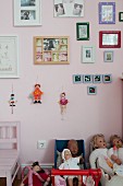 Dolls in dolls' pram below framed pictures and marionettes on pink-painted wall