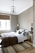 Bedroom in muted natural shades with many scatter cushions on bed, modern chandelier and antique-style bedside tables