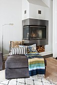 Scatter cushions and blanket in shades of blue and green on ottoman; modern fireplace with L-shaped glass door in background