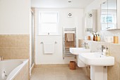 Pale bathroom with twin sinks on projecting wall below mirrored cabinets and sand-coloured tiles on floor and walls