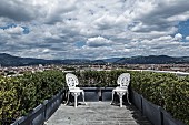 Two white, vintage, Rococo-style metal chairs positioned in corners of planted roof terrace with magnificent panoramic view