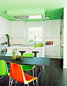 Colourful plastic chairs at black table, dark wooden floor, white modern counter and fitted kitchen in background below green-painted ceiling in open-plan interior