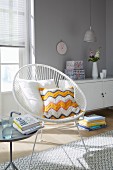 A cushion with a crocheted cover on a white retro chair in front of a sideboard on a light grey wall