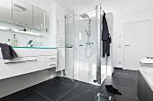A long wash stand built into a white unit with a multi-door, mirrored cabinet and an illuminated glass shelf above it and a glass shower cubicle in the background in modern, black-and-white bathroom with large black floor tiles