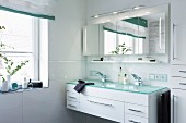 A double wash basin with a turquoise glass platter over a white cupboard with an illuminated, four-door mirrored cabinet
