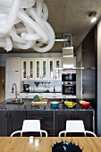 View across dining table to counter below extractor hood suspended from concrete ceiling; pendant lampshade made from plastic hoses in foreground