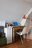 Small, eclectic workspace under sloping ceiling with antique table, shell chair, modern filing cabinet and stack of old trunks