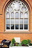Neogotisches Fenster mit Rosetten Elementen in Ziegelfassade einer umgebauten Kirche, davor Hochbeet und Aussengrill auf Terrasse