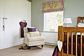 Foot of bed, pale grey armchair and white wardrobe in corner of pale-grey bedroom with patterned roller blind on window