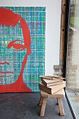Books on wooden stool in front of large, modern portrait of man in artist's studio