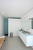 Designerbad mit weißem Waschtischmöbel und zwei Waschbecken, langer Spiegel mit Wandbeleuchtung, im Hintergrund weiße Wand vor Duschbereich