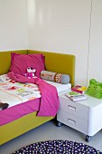 Comfortable bed with lime green upholstered frame and white cabinet on castors as bedside table in child's bedroom
