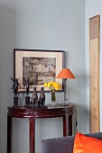 Sculptures and delicate table lamp on antique, semicircular console table against pale grey wall