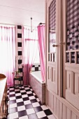 Vintage bathroom with chequered floor and wall, pink curtain on window and white-painted fitted cupboards to one side
