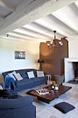 Schlichter Bodentisch aus Holz und blaue Sofagarnitur in mediterranem Wohnraum mit weisser Holzbalkendecke