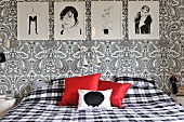 Bedroom with black and white drawings, floral Paisley wallpaper, gingham bed linen and red scatter cushions providing a splash of colour