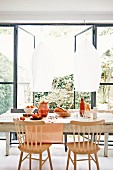 Simple wooden chairs around fruit and bread on table below pendant lamps with white lampshades; open terrace doors with view into garden