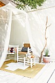 Translucent mosquito net hanging from tree, stool, various chairs and sisal rug on white-painted wooden terrace
