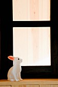 weiße Hasenfigur auf Holzfenstersims vor schwarzem Fensterrahmen