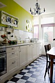Kitchen counter with white cupboards against wall painted green with tiled splashback; chequered floor tiles