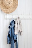 Straw hat and denim jacket hanging from coat pegs on white wood panelling