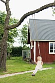 Little girl playing on swing hung from tree in front of wooden house in summery garden