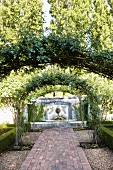 Climbers on arched trellising over paved path leading to fountain on wall in summery garden
