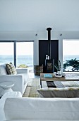 White sofa set around delicate coffee table, wood-burning stove flanked by French windows in contemporary living room with panoramic view of ocean