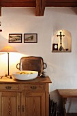 White fruit bowl and table lamp on wooden sideboard against wall next to shrine in small, illuminated niche
