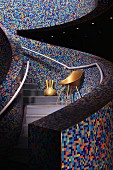 Curved staircase with balustrade walls covered in colourful mosaic tiles; gold-painted chair and vase under silver-grey handrail