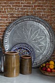 Ceramic pots with olive-green glaze, plate with ethnic pattern and large, pewter platter in front of brick wall
