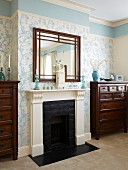 Mirror above fireplace with white surround flanked by chests of drawers in room with floral wallpaper and blue frieze