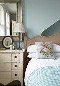 Floral cushion on country-house bed next to chest of drawers and mirror on pale blue wall