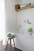 Toiletries and towels on wooden stool next to bathtub with white-tiled surround in corner of bathroom
