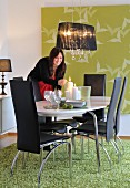 Pendant lamp with black fabric lampshade over black leather chairs and oval dining table on rug, large artwork with bird motif on wall and woman lighting candles